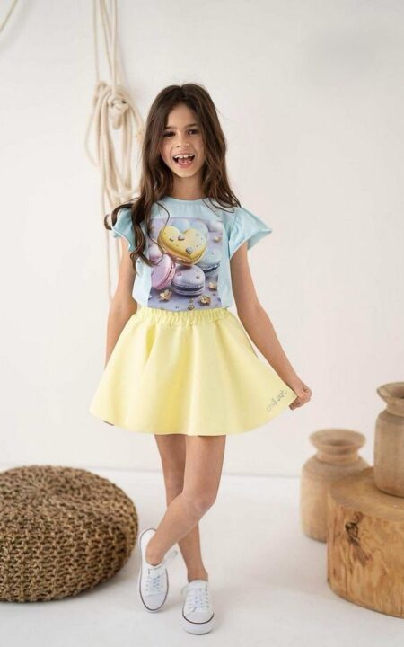 Miętowa bluzka dla dziewczynki z kolorowym nadrukiem ciastek, zestawiona z żółtą spódniczką.