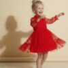 Młodsza dziewczynka w czerwonej sukience tiulowej z długimi rękawami, uśmiechająca się i tańcząca.