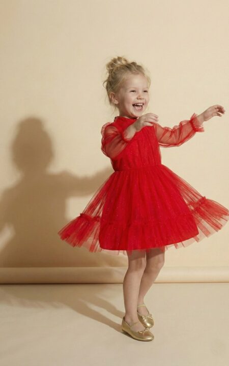 Młodsza dziewczynka w czerwonej sukience tiulowej z długimi rękawami, uśmiechająca się i tańcząca.