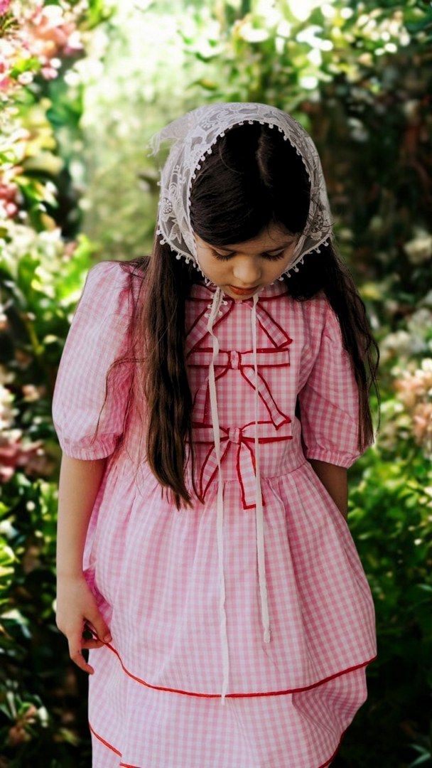 Różowa sukienka w kratkę dla dziewczynki z białą koronkową chustką na głowie. Idealna na letnie przyjęcia i rodzinne uroczystości.