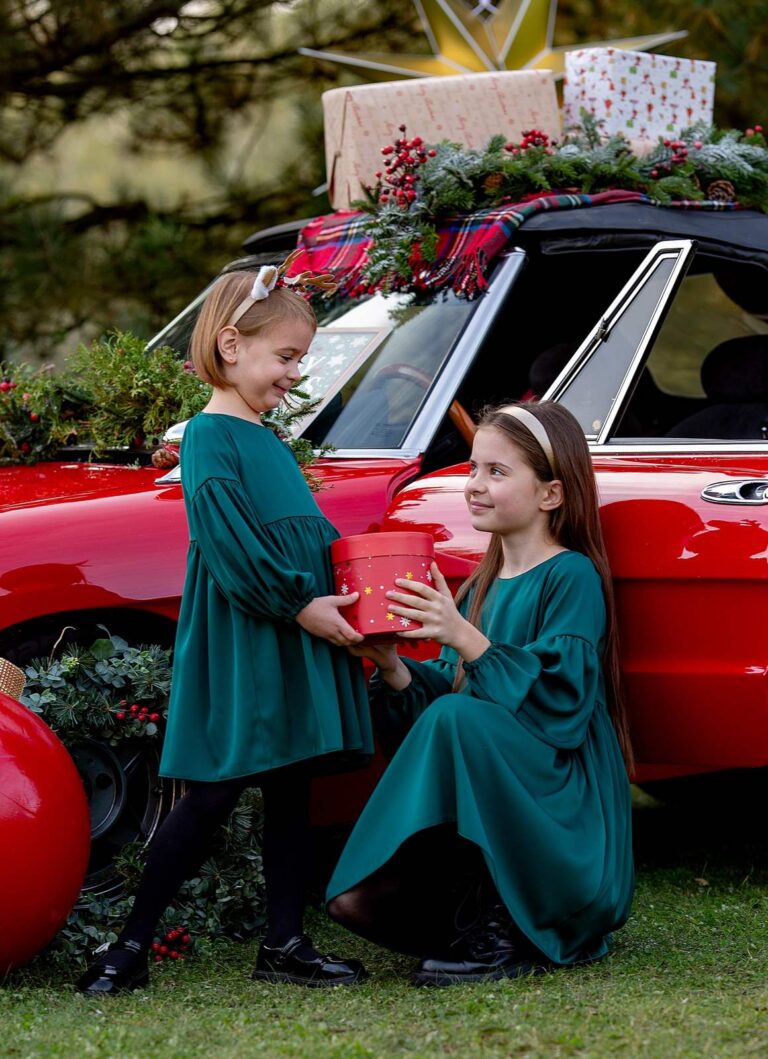 Satynowa sukienka w kolorze butelkowej zieleni dla dziewczynki, idealna na święta, prezentująca dwie dziewczynki w radosnej scenerii.
