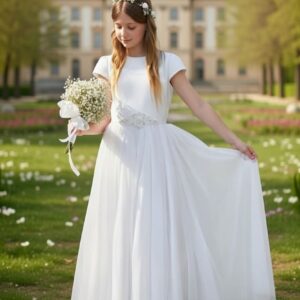 Biała długa suknia dla dziewczynki na komunię z ozdobnym paskiem i bukietem kwiatów w dłoni.