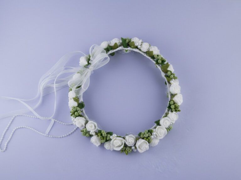 Tradycyjne wianek na komunię z białymi różami i zielonymi dodatkami, ozdobiony wstążką.