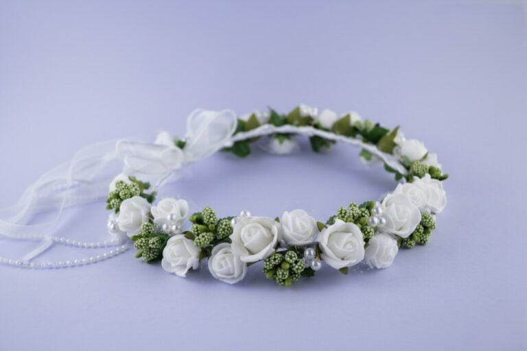 Wianek z białymi różami i zielonymi dodatkami, ozdobiony perłami i wstążką. Idealny na uroczystości i specjalne okazje.